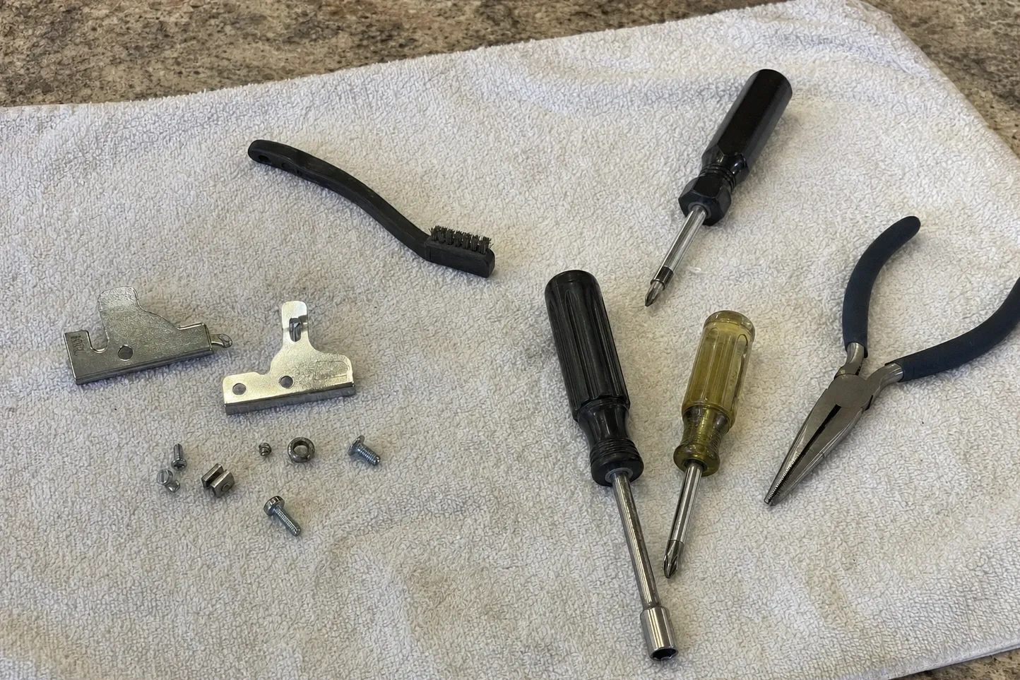 Two steel brackets and their screws set out on a towel with a wire brush, nut driver, screwdrivers and needle-nose pliers